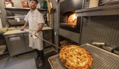 Restaurant executive chef Christopher Cameron, holds a pizza paddle with a cooked pizza on it, with a glowing wood-fired pizza oven to his right.