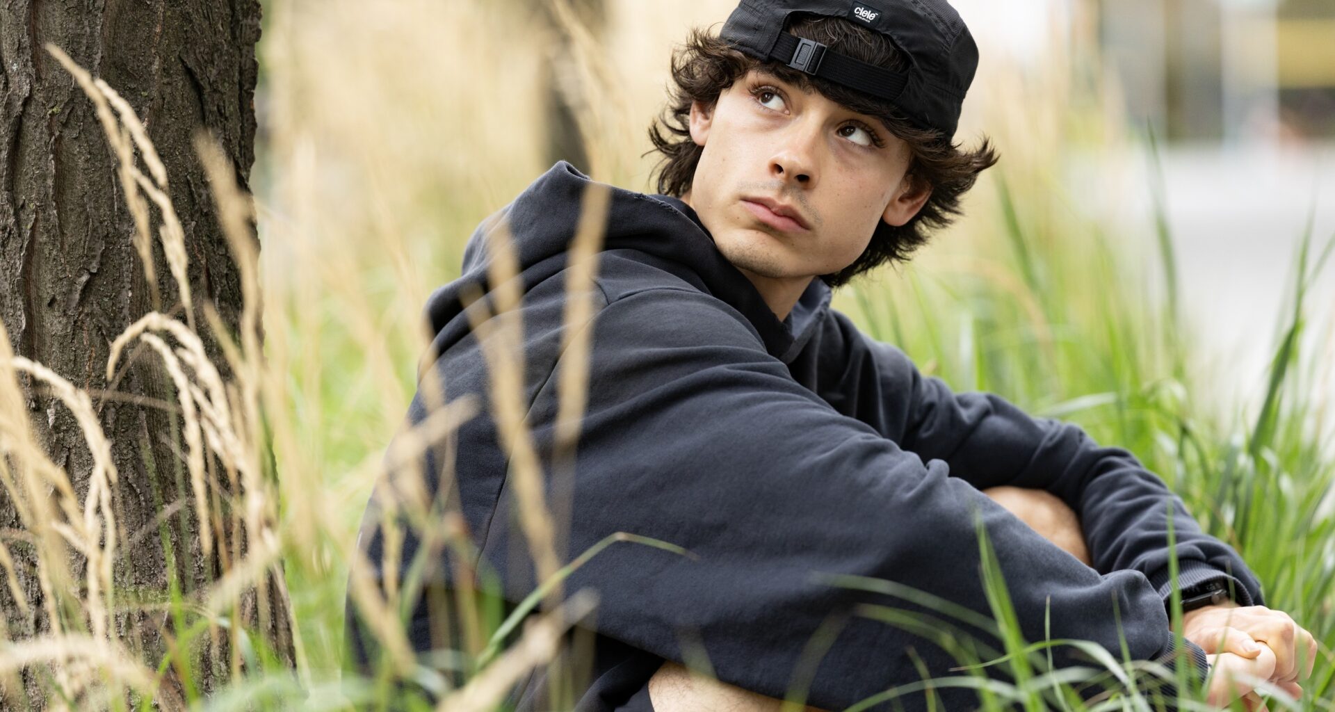 Jacob Whiteduck-Lavoie sitting in the grass near a tree, wearing a backwards ball cap.