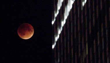A reddish, eclipsed moon next to an office building.