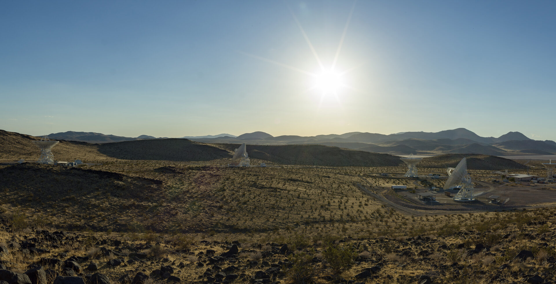 A wide desert landscape featuring several large white satellite dishes pointing toward a bright sun shining in a clear blue sky above distant mountain ranges.