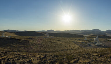 A wide desert landscape featuring several large white satellite dishes pointing toward a bright sun shining in a clear blue sky above distant mountain ranges.