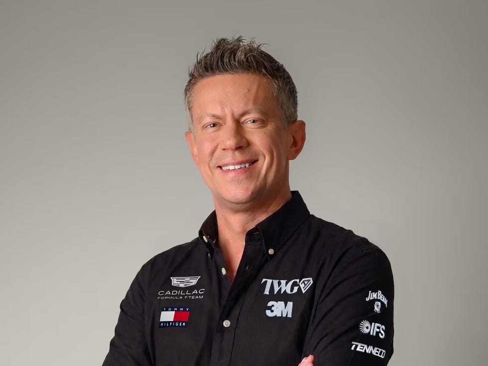 Smiling man in a black shirt with racing team logos, posing with arms crossed against a neutral background.