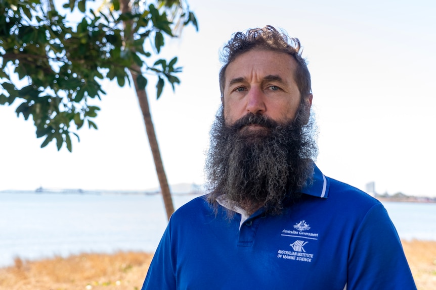 A man with a big beard and blue polo shirt standing with the ocean behind him