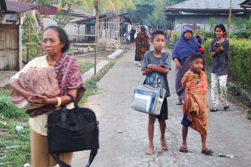Women and children carrying bags and walking along a road past houses, looking anxious.