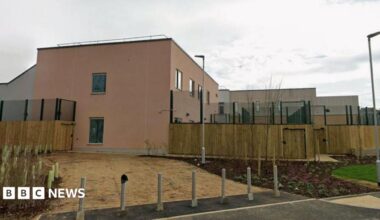 The image shows the outside of a mental health trust: wooden beams and green gates can be seen on the exterior and the outside of the building is painted a creamy pink.