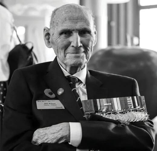 A man wearing a naval uniform with a poppy and medals smiles at the camera.