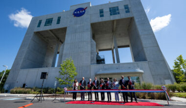 A group of people in suits stand in front of a massive square building that has two big square openings in it that go all the way through. The people all hold big pairs of scissors, as they wait to cut a red, white, and blue ribbon placed in front of them.