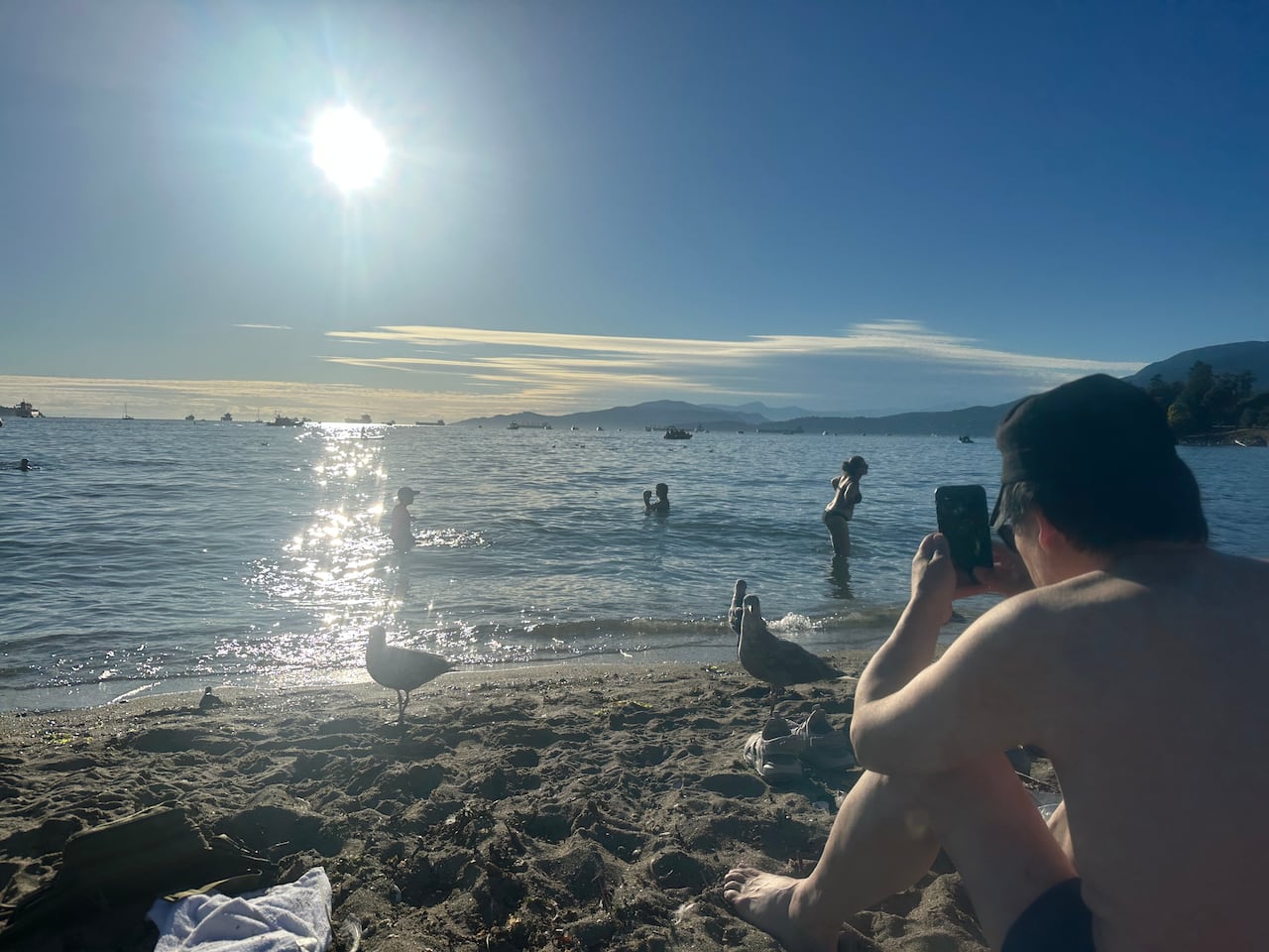 A person is pictured holding their phone toward the horizon at English Bay, midday, for a photograph.