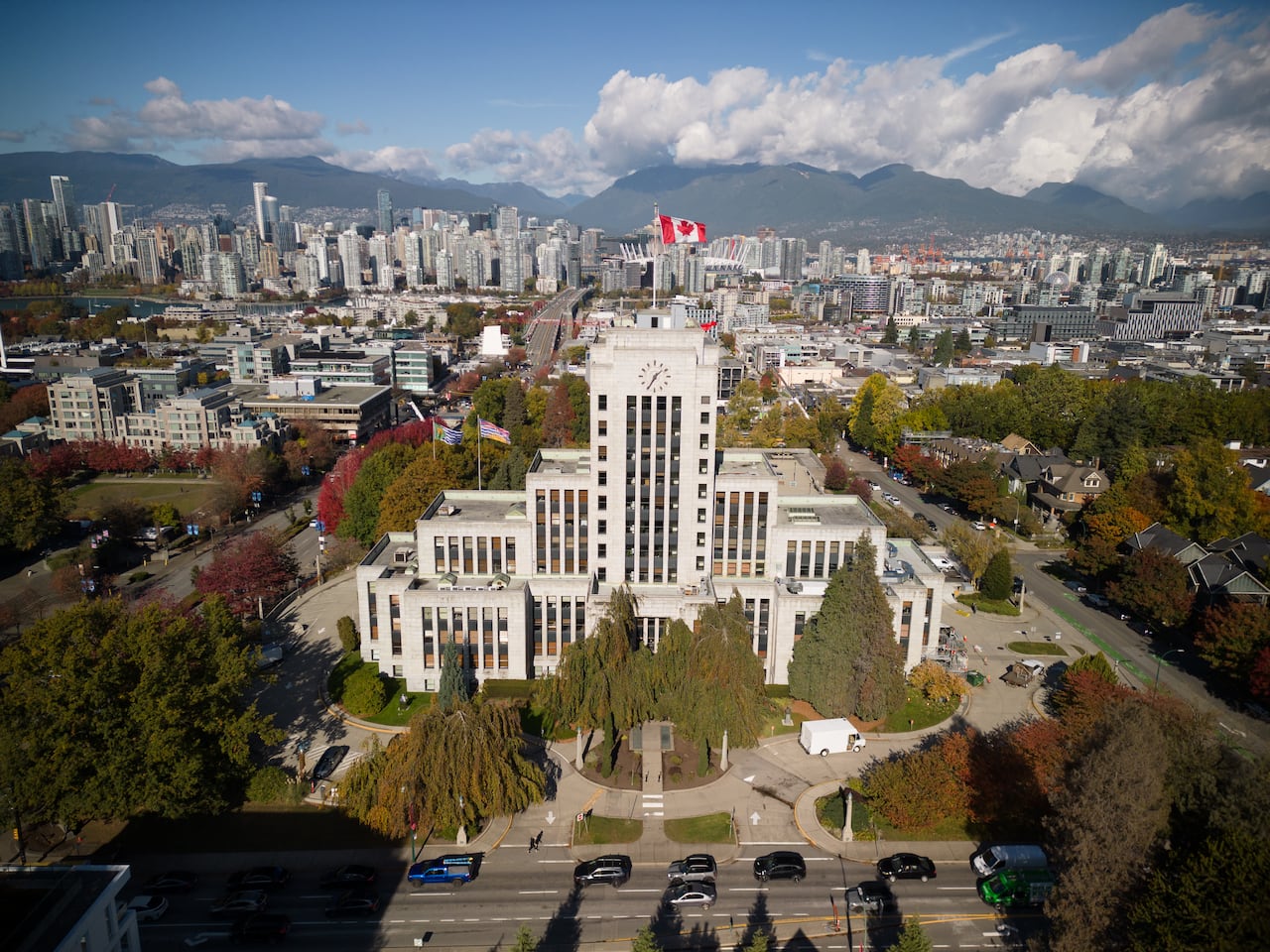 An aerial photograph of Vancouver City Hall with a Canada flag waving on top and in the background the highrise towers of Vancouver and North Shore mountains rise up.