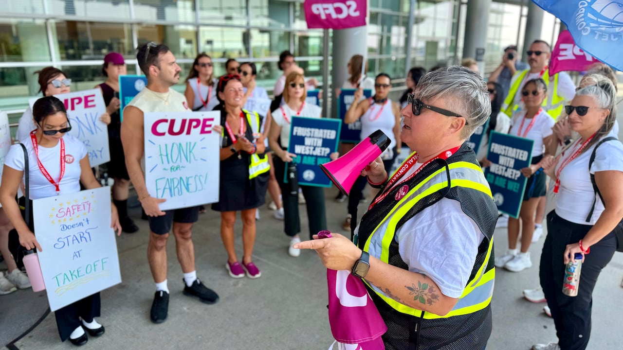 A person with short grey hair wearing a bright yellow vest speaks into a small pink megaphone as people holding signs surround them