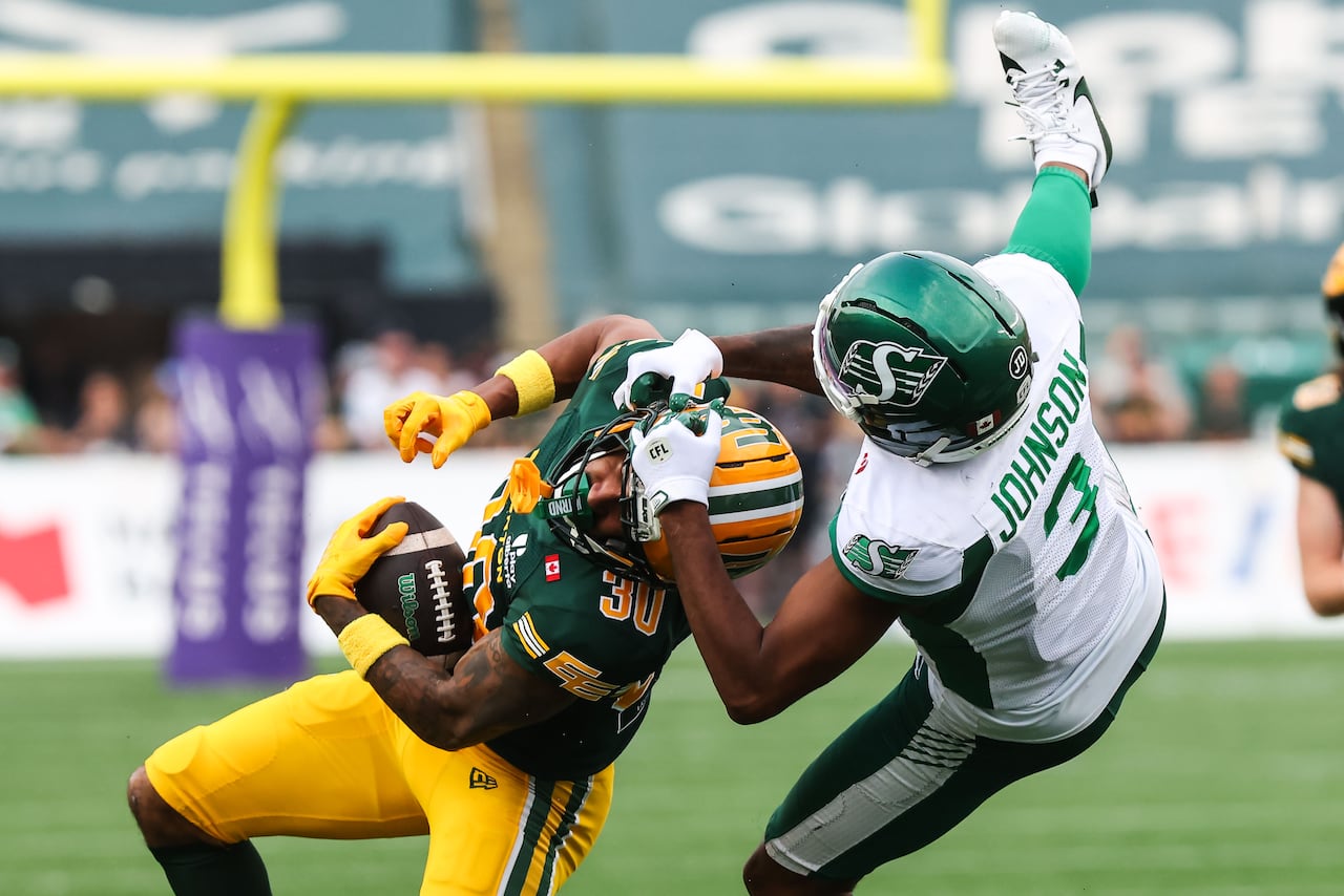 Football player wearing a green and white jersey holds onto the helmet of a football player wearing a green and yellow jersey, pulling him to the ground.