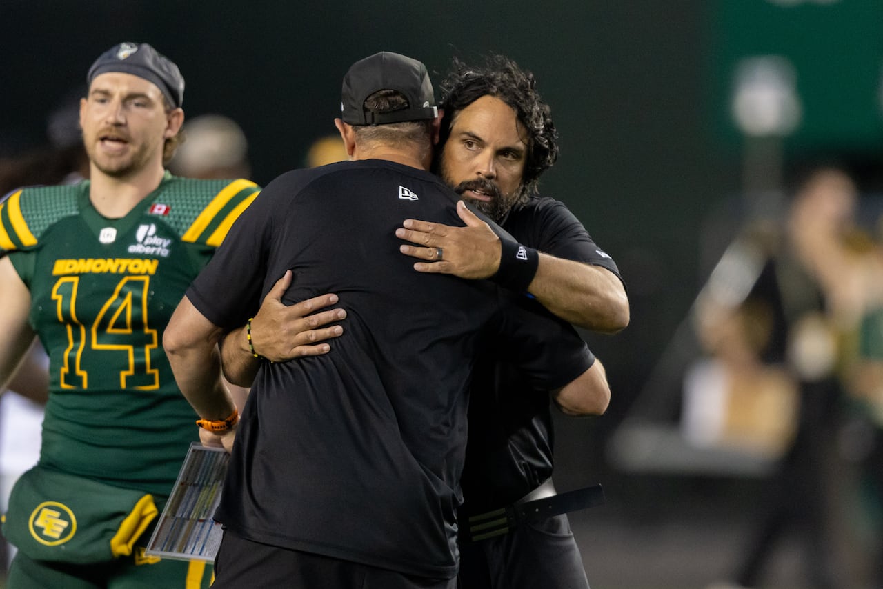 Two men with brown skin, wearing black shirts, hug on a football field. Players pass by around them.