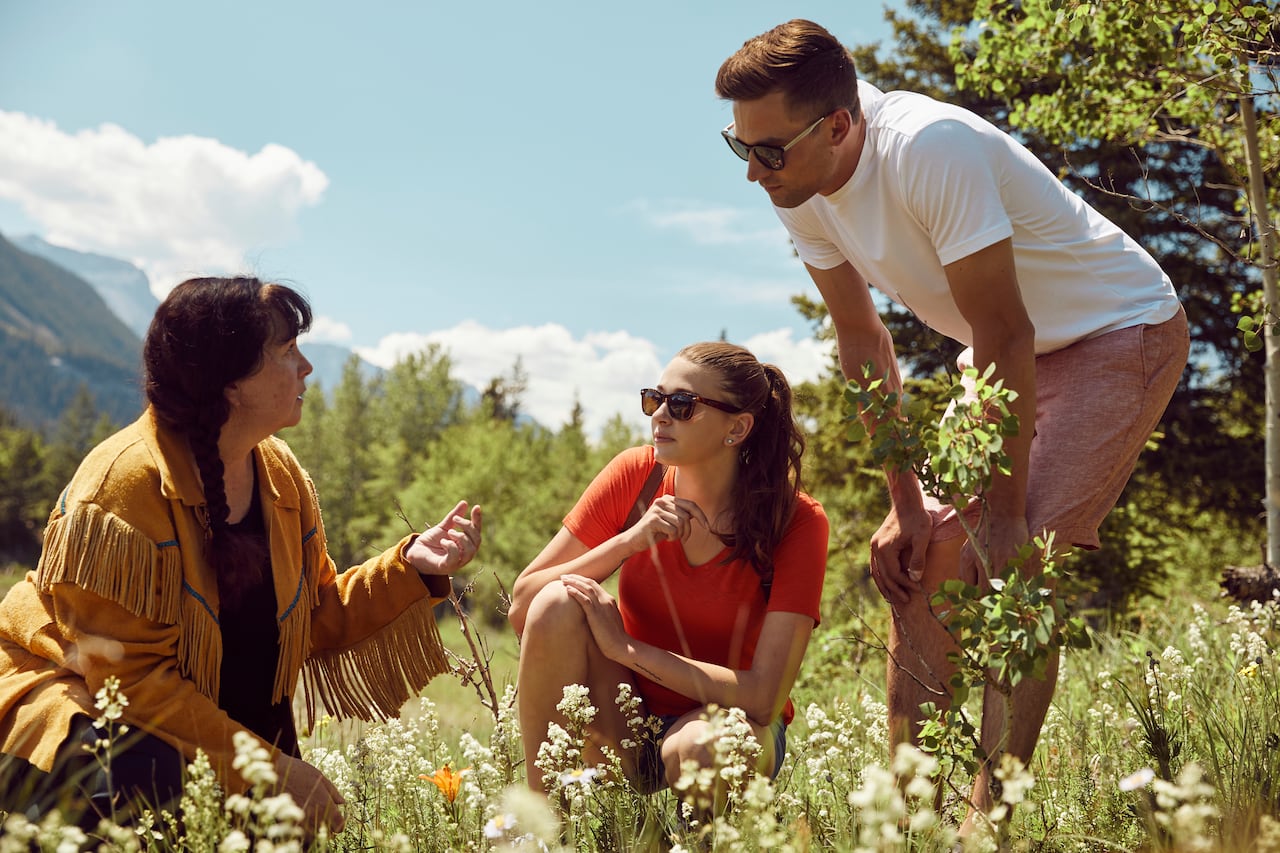 A woman to the left is kneeling in a meadow of flowers. Two other people are looking at plants.