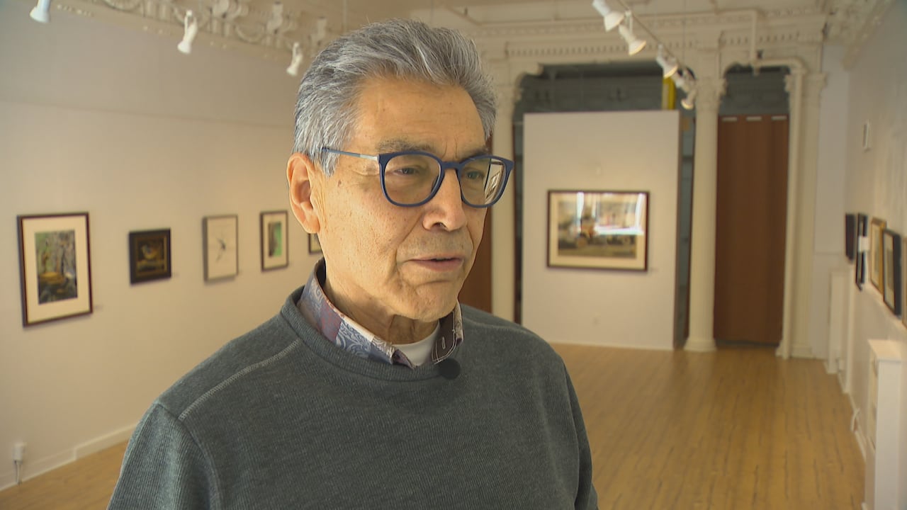 A man wearing glasses, a sweater and a collared shirt stands in an art gallery with paintings hanging on the walls.