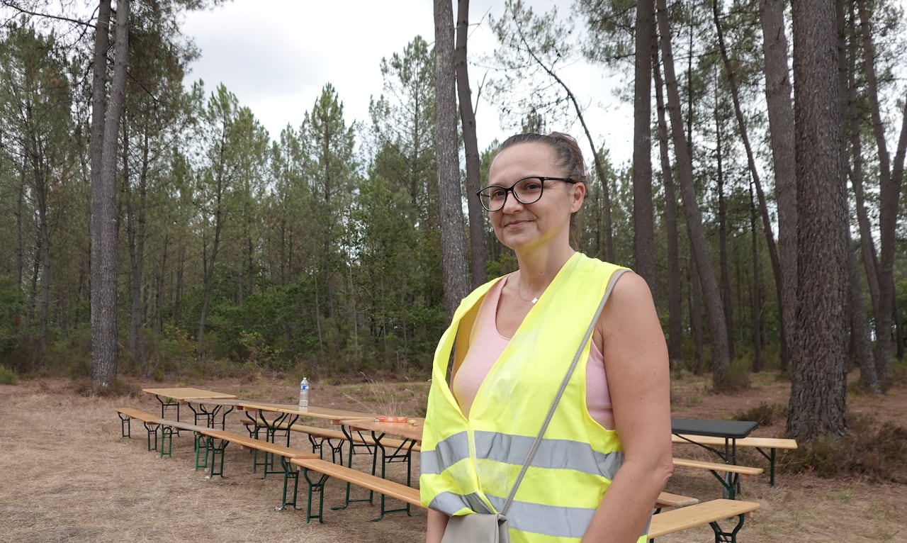 A woman with a high viz vest stands in front of a table under tall pine trees.