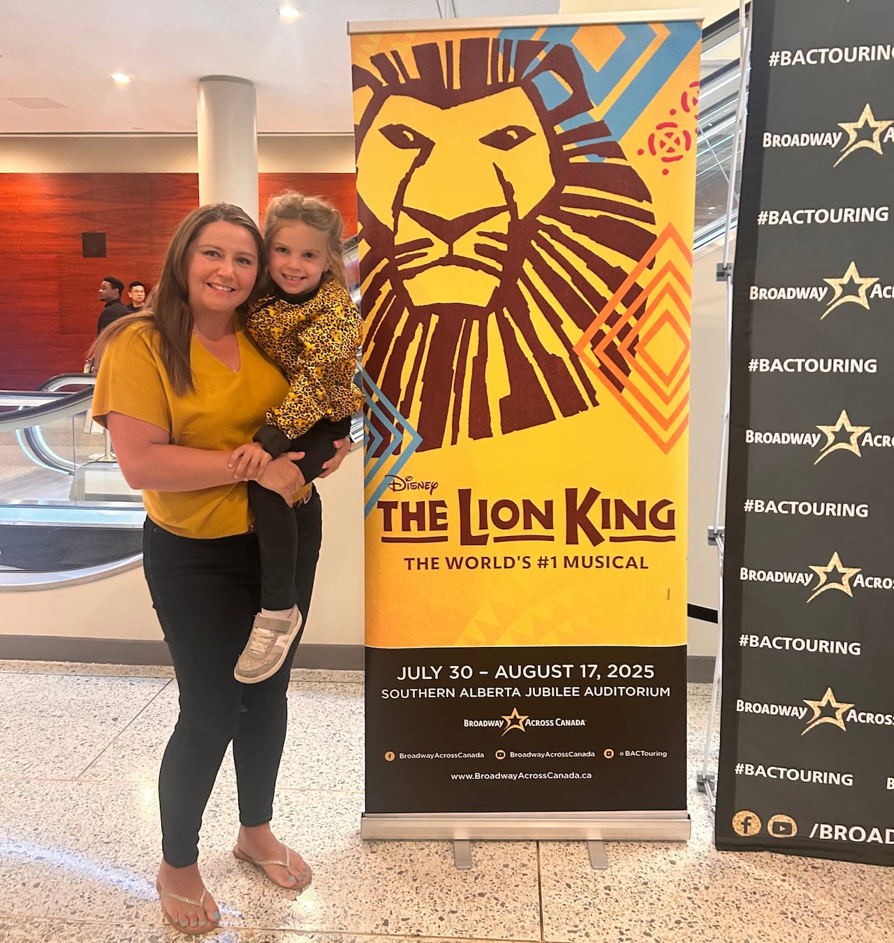 A mom holds her daughter on her hip in front of a sign for the Lion King musical