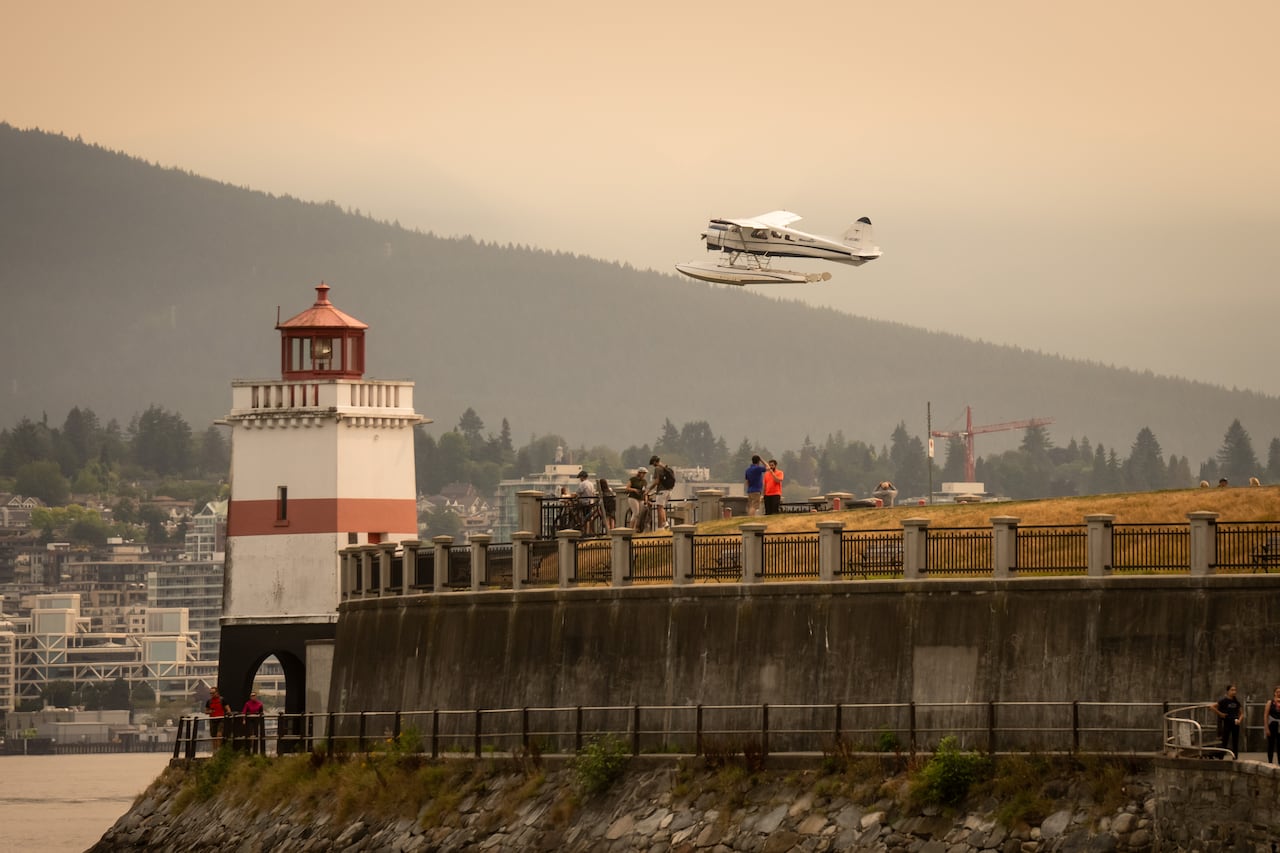 A seaplane takes off over a lighthouse amid visibly orange skies.