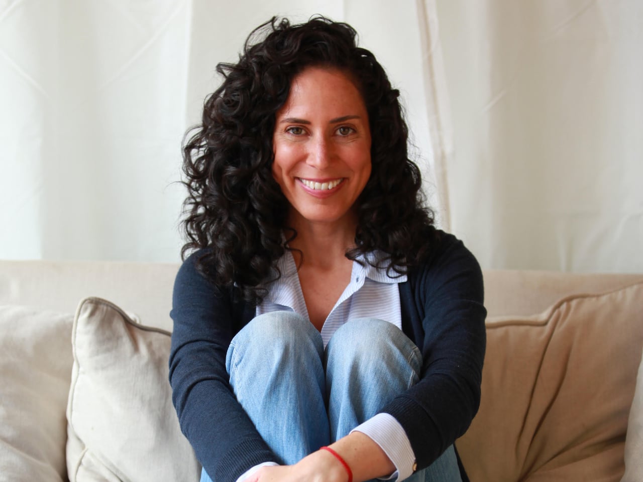 A woman with dark, curly hair, a navy shirt and jeans sits on a beige couch smiling at the camera.
