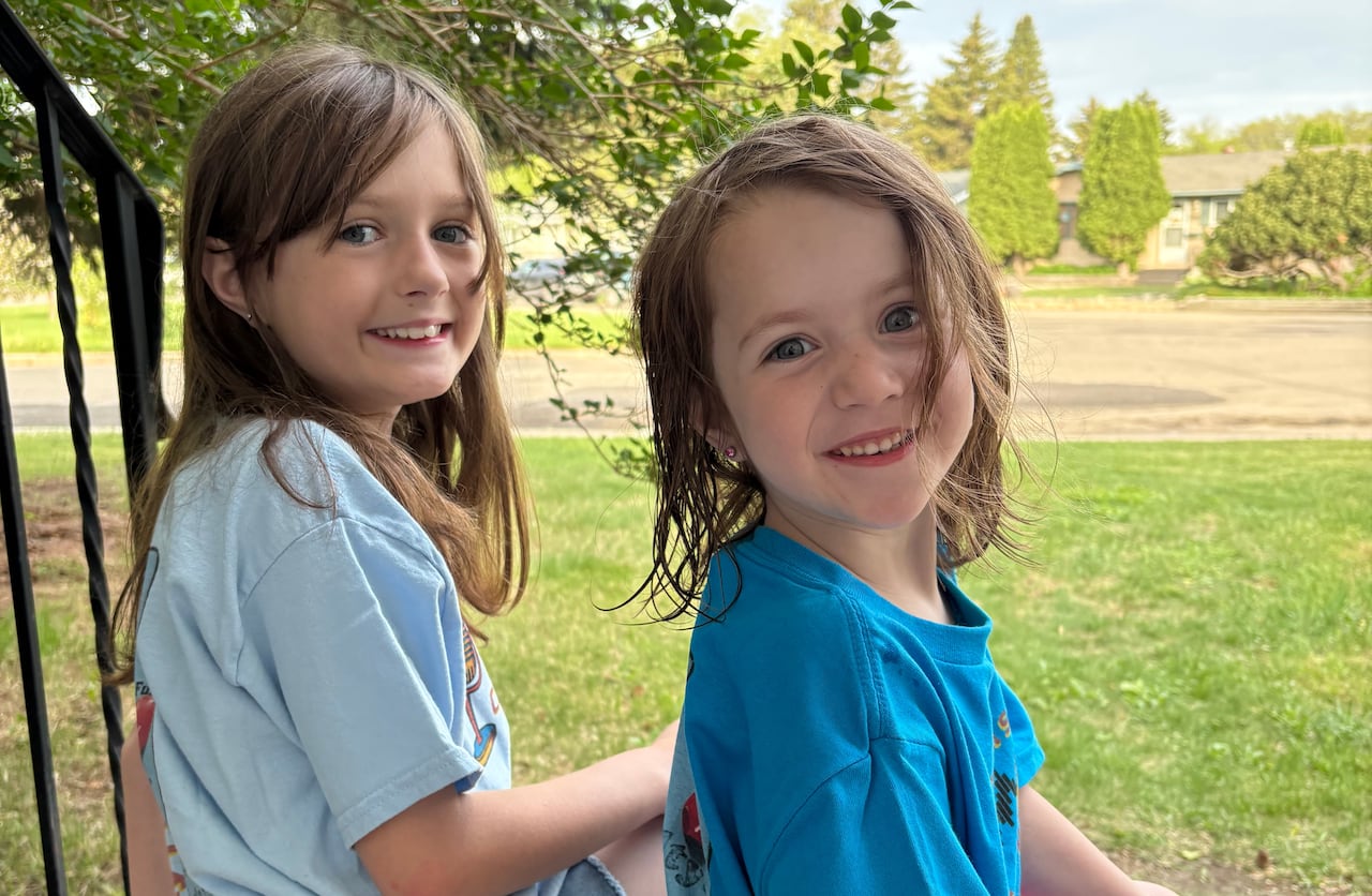 Two little girls sit on the top step of a porch with a park in the background.
