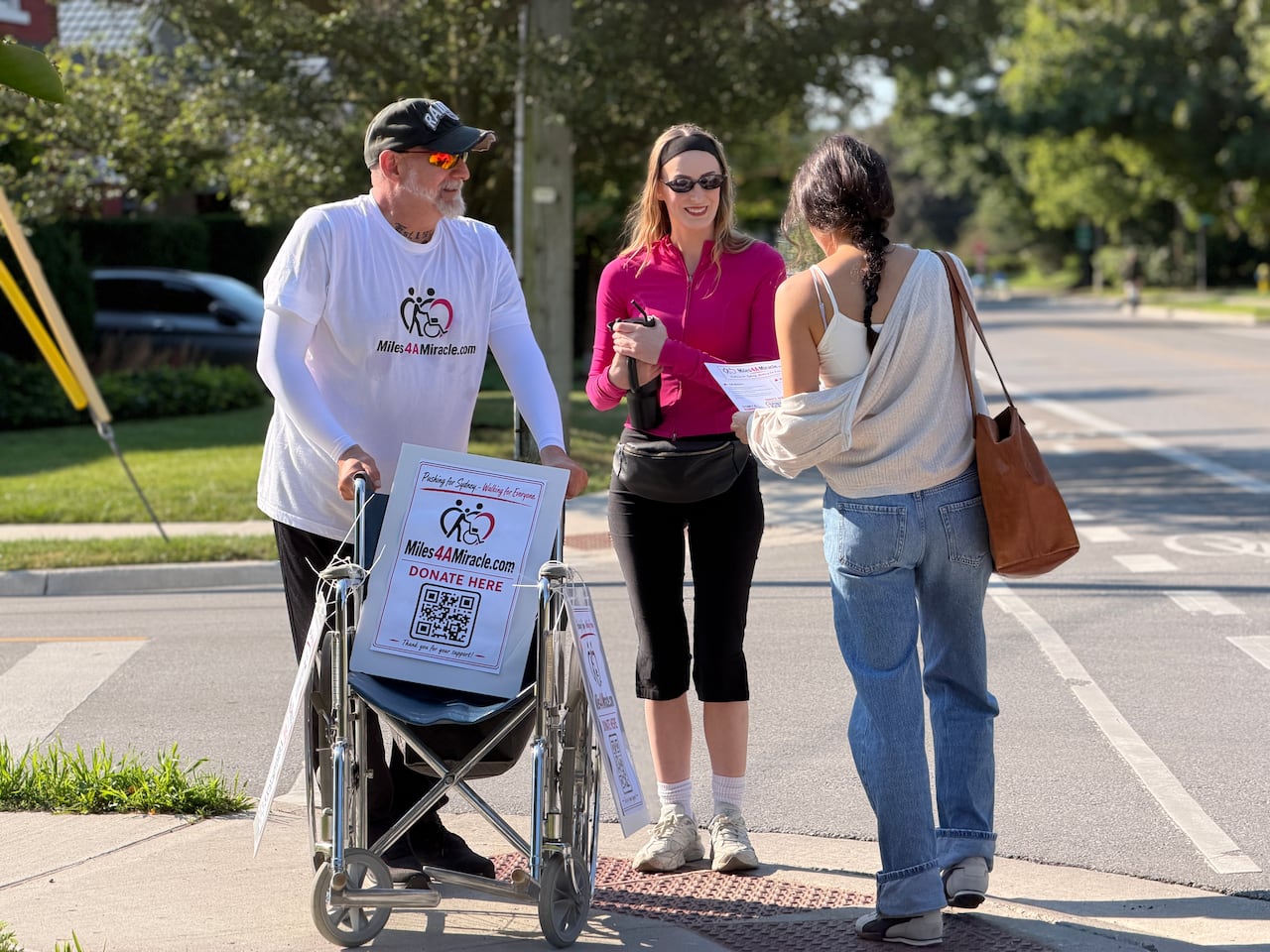 A man pushing a wheelchair speaks with a woman reading a flyer. Another woman smiles
