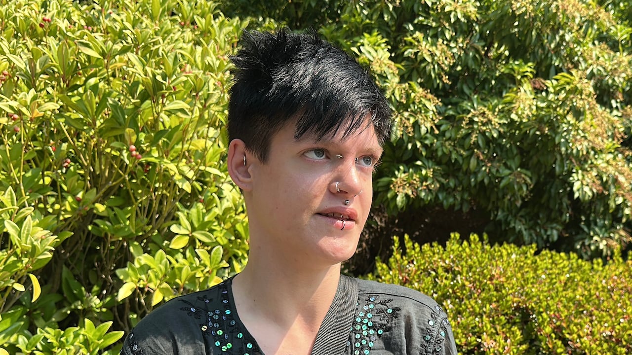 A person with black hair, piercings on eyebrows and face, wearing a black shirt stands in front of some bushes as they are interviewed.