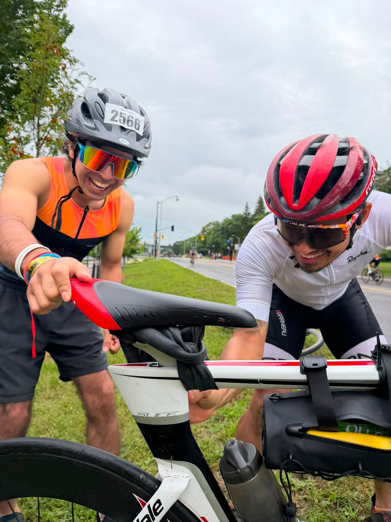 Two men wearing biking gear next to a bike seat that has an old inner tube wrapped around it to make it secure.