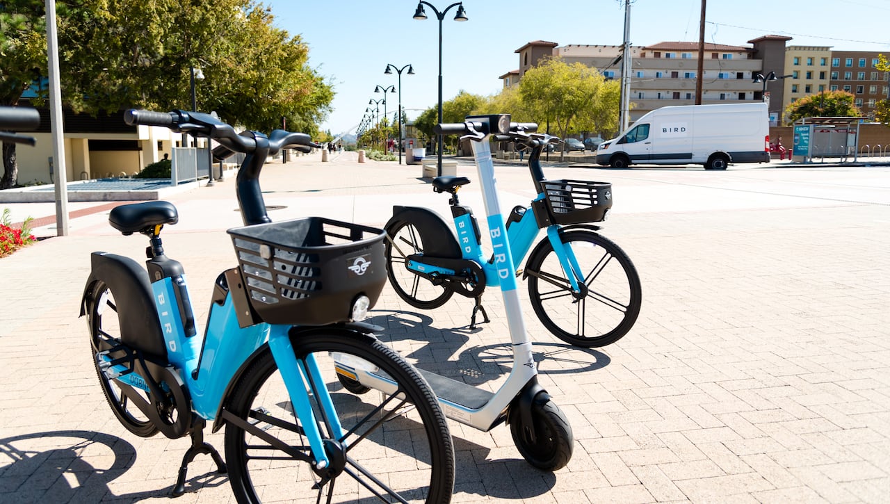 Two blue and silver e-bikes are parked on a cobblestone street with a silver scooter between them