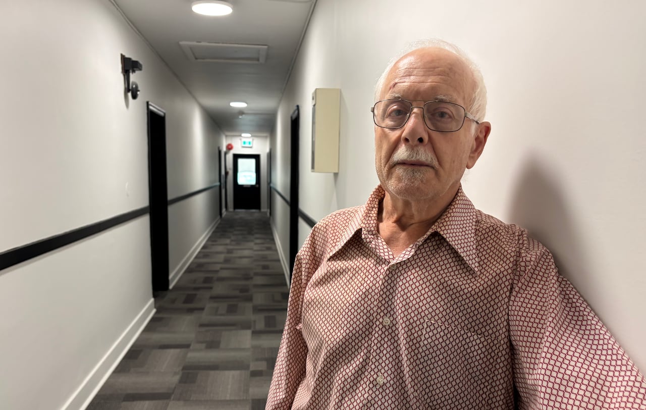 A man in a red and white collared shirt stands leans along a wall in a hallway.