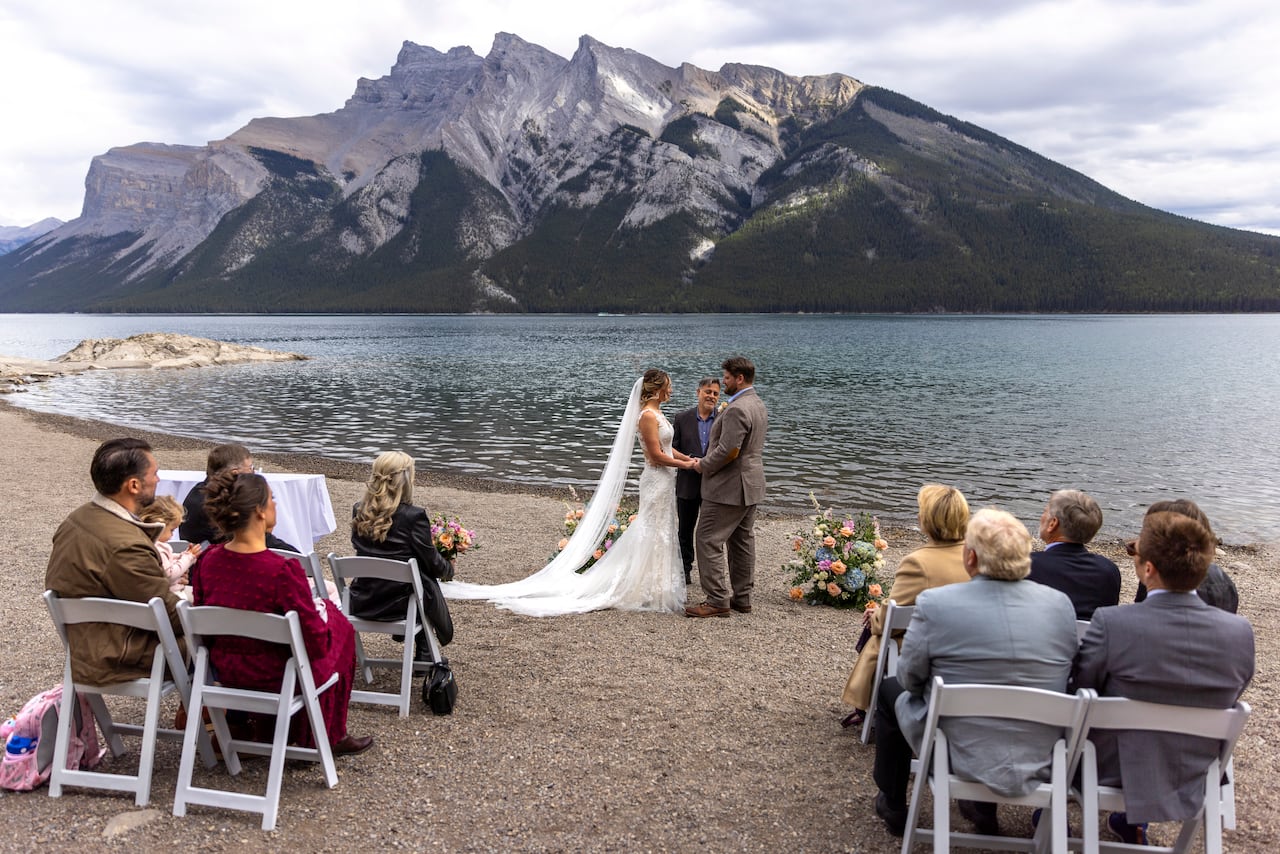 A man and woman getting married in front of a lake with mountains in the background. 9 people are sitting in chairs, watching the ceremony.