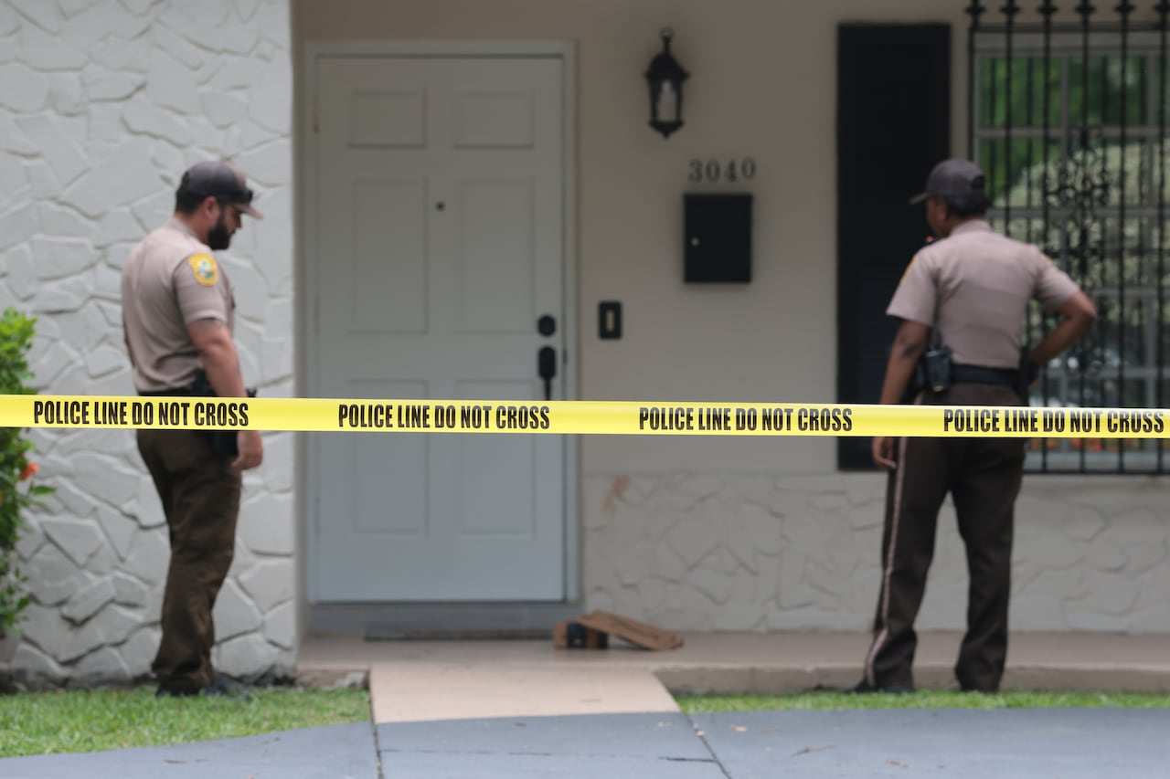 Police men stand outside a door, a line of caution tape up reading : Police Line do not cross