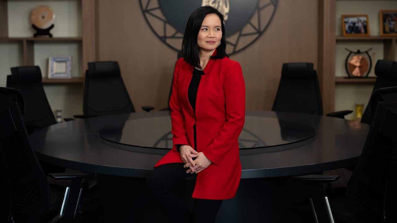 A woman in a red jacket looks away from the camera while sitting on a table.