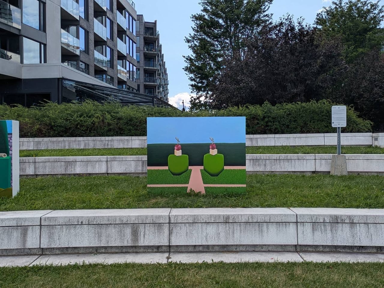 A print of a surreal painting of a minimalist garden landscape is installed on a grass lawn. A highrise appears in the background.