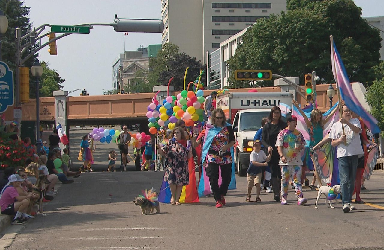 A parade of people dressed in bright colours with various Pride flags, and trans flags, with a small dog on a leash dressed in a rainbow tutu. A U-Haul truck is behind them, hauling a parade float with rainbow balloons.