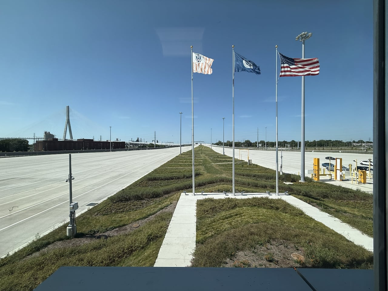 Three flags are seen in a grassy median, with large expanses of road on either side, and a bridge in the background.