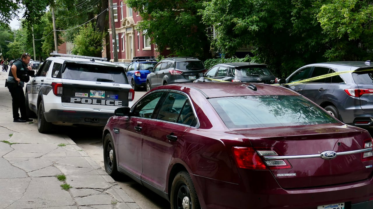 A series of parked cars surround on a city street includes a police vehicle with an officer standing outside it. Police tape lines a section of the street