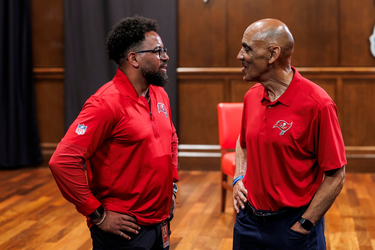 Two Black men wearing red Tampa Bay Buccaneers merchandise have a conversation.