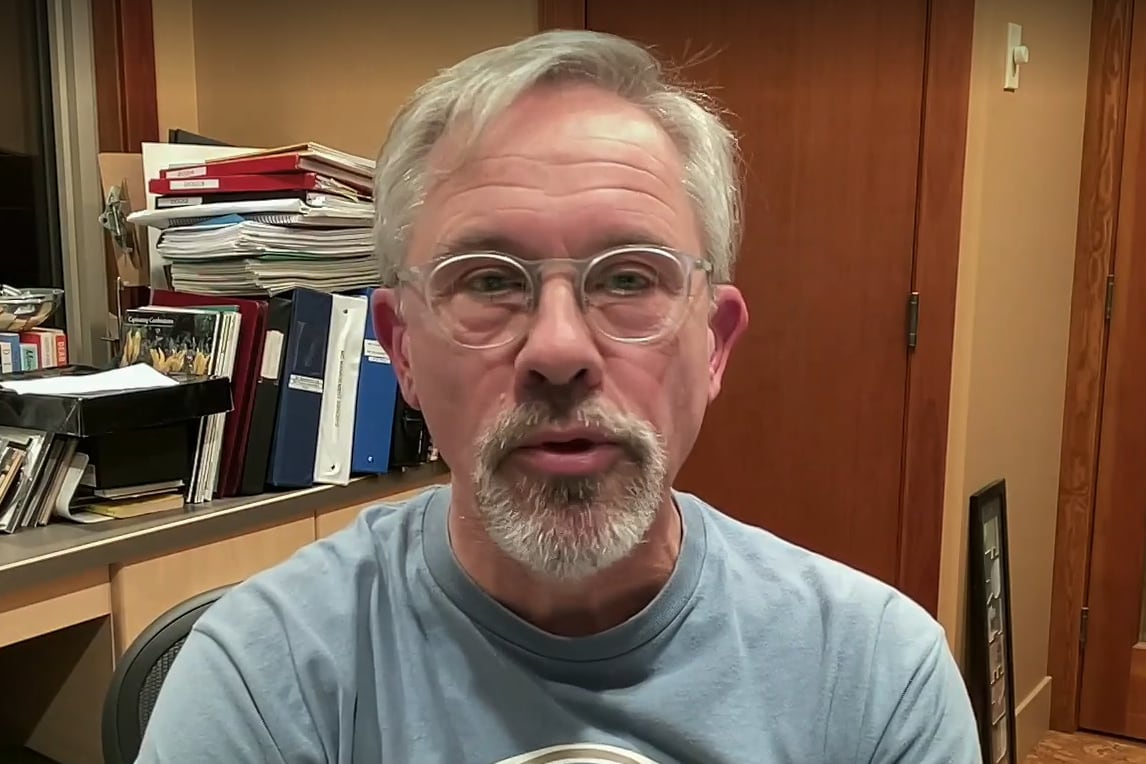 A man wearing glasses. There is a stack of books behind him