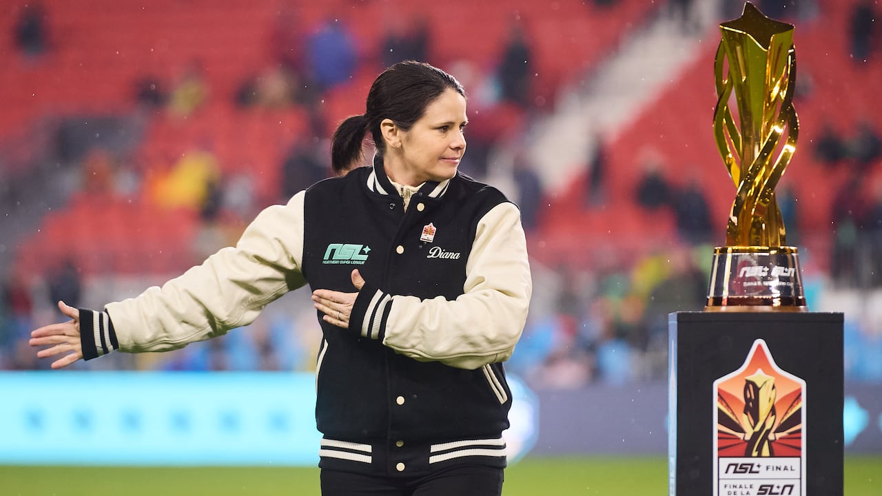 A woman in a letterman style jacket stands next to a gold trophy on a soccer field.
