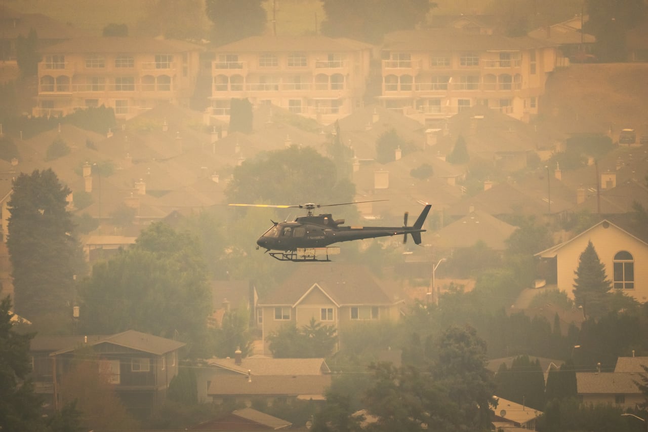 A black helicopter above a residential area shrouded in orange-brown smoke.
