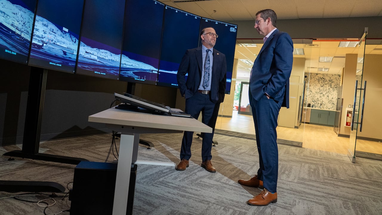 Two men talk with each other in a room with a line of monitors showing a large infrared image