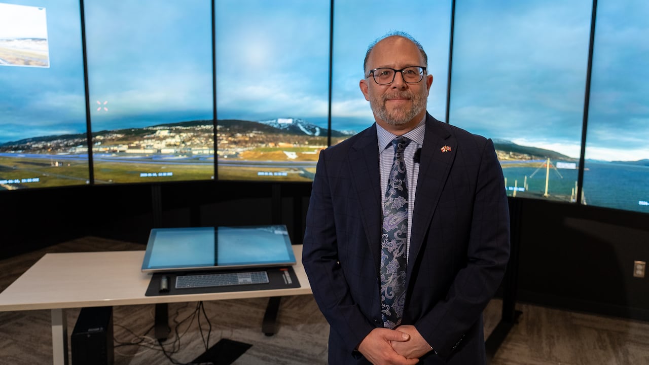Man in glasses stands in front of a row of monitors showing an image of an airport with water around it.