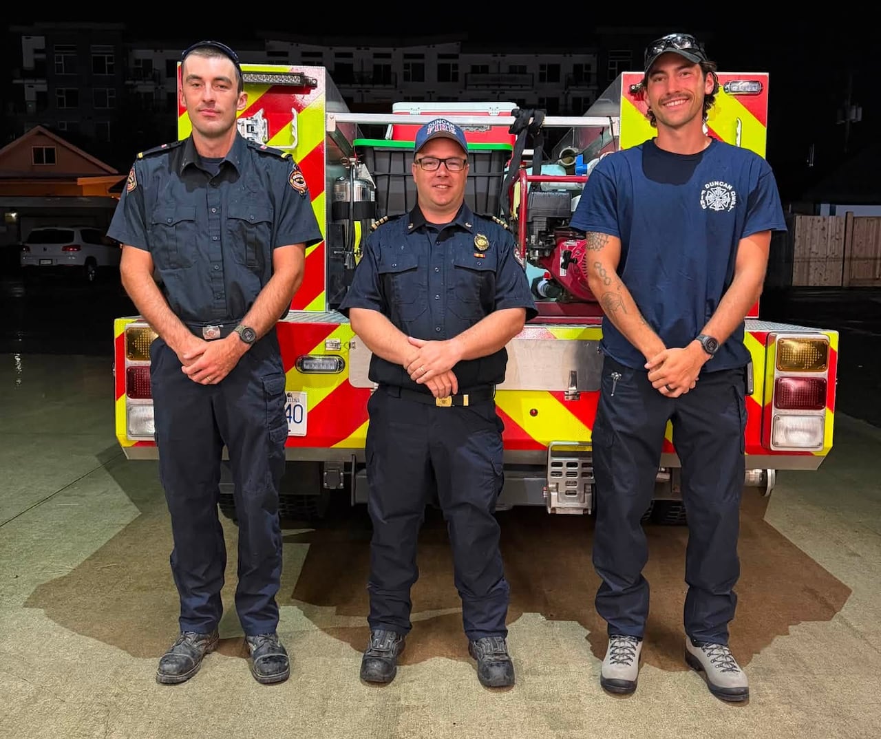 Three male firefighters stand in front of a firetruck.