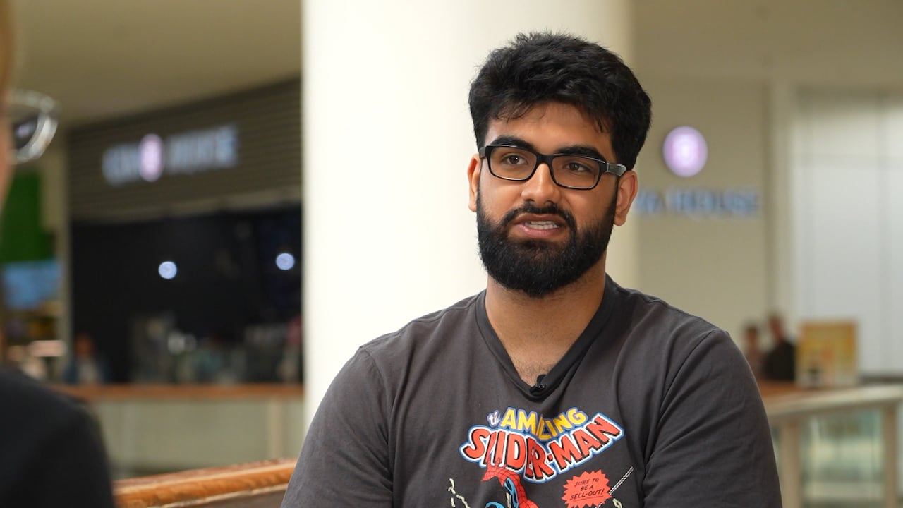A man wearing a superman shirt sits on a chair in the middle of a mall.