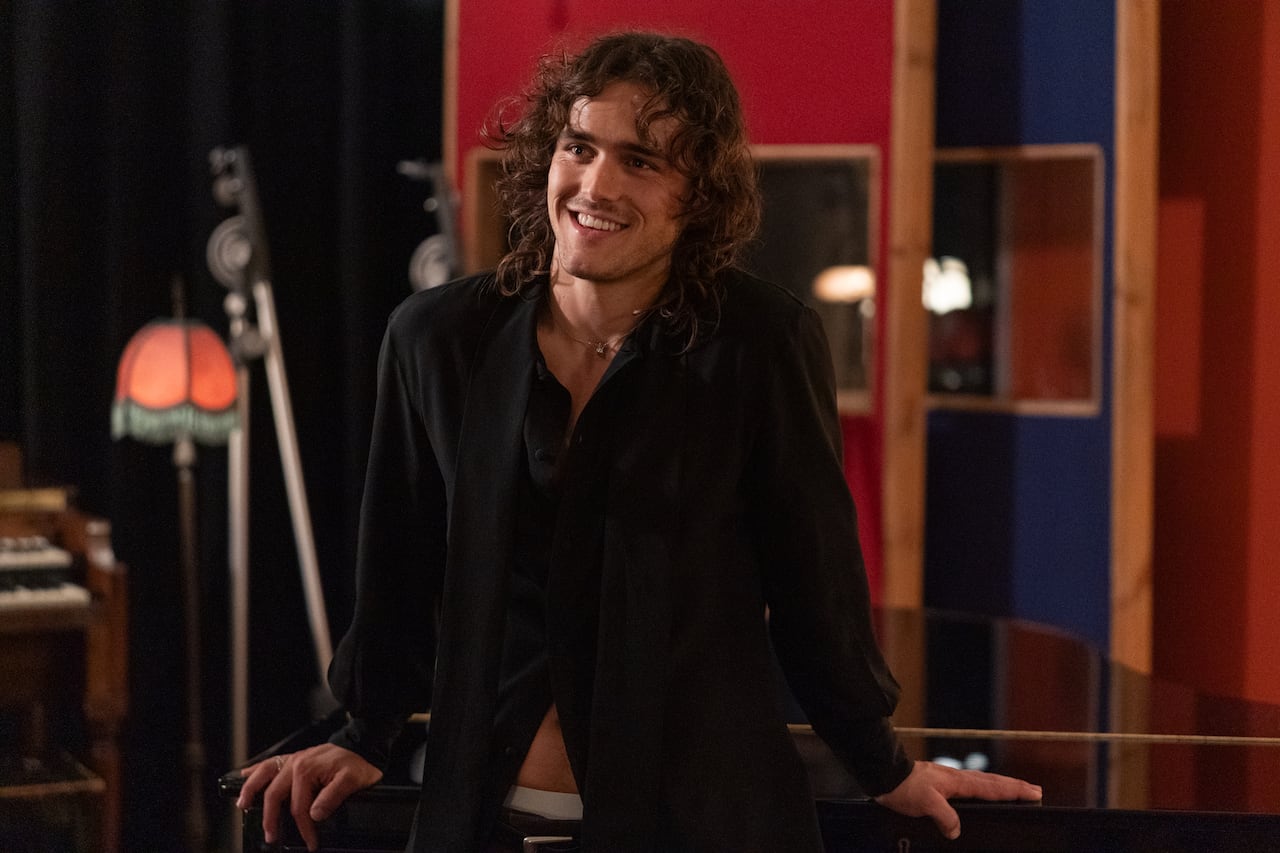 A man with long hair leans back against a piano, smiling.