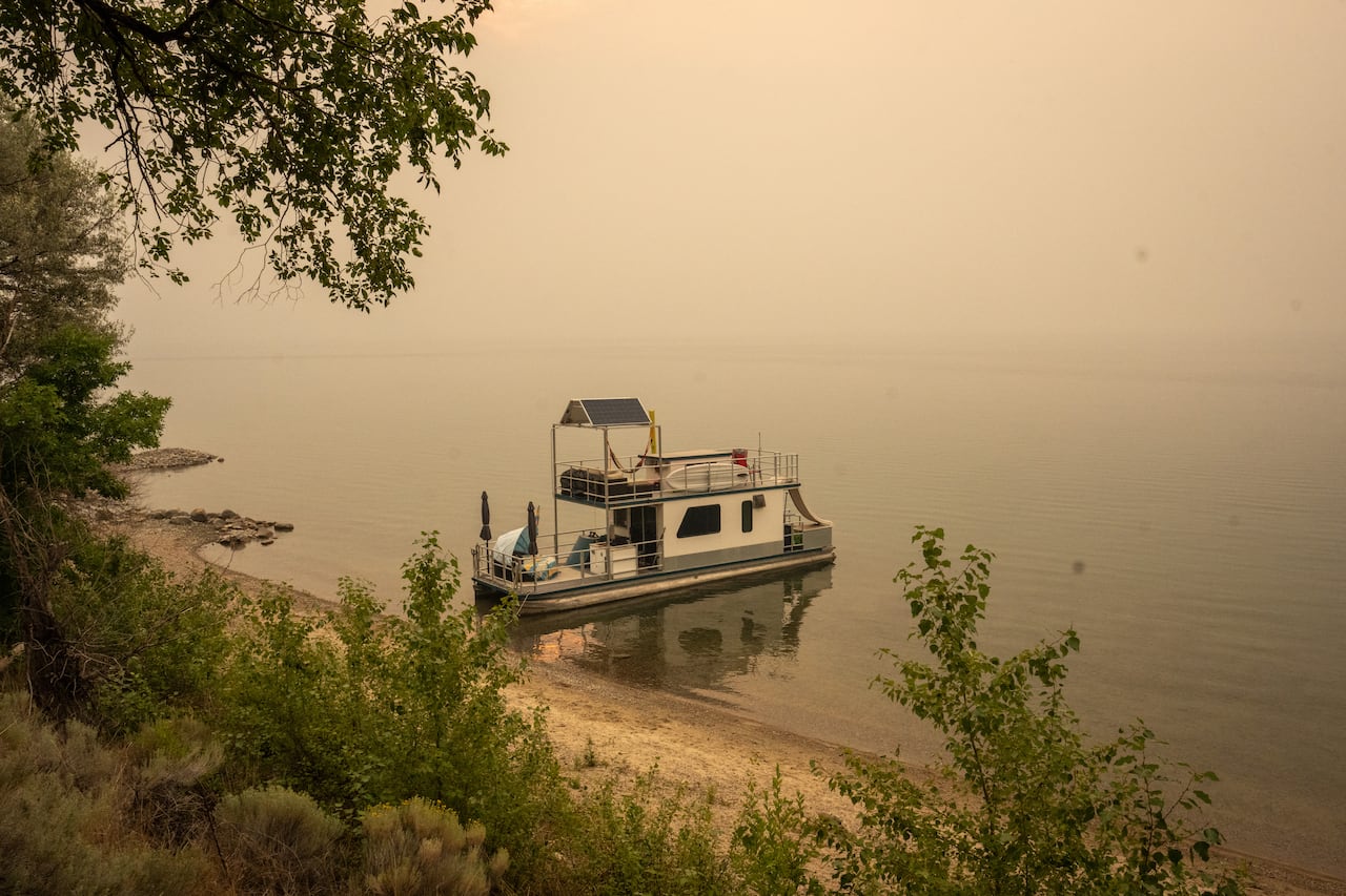 A houseboat moored at a beach on a still lake with the horizon obscured by grey-brown smoke.