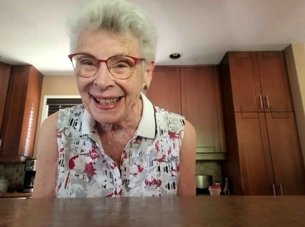 An elderly woman wearing glasses smiles in a kitchen.