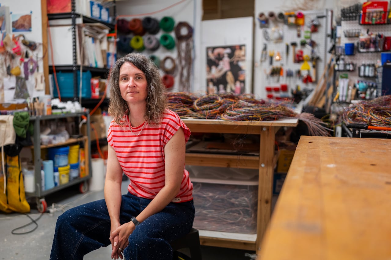 Emily Hermant, in a red and pink striped t-shirt and jeans, with medium length brown hair, sits in a studio full of spools of telephone wire and ethernet cable.