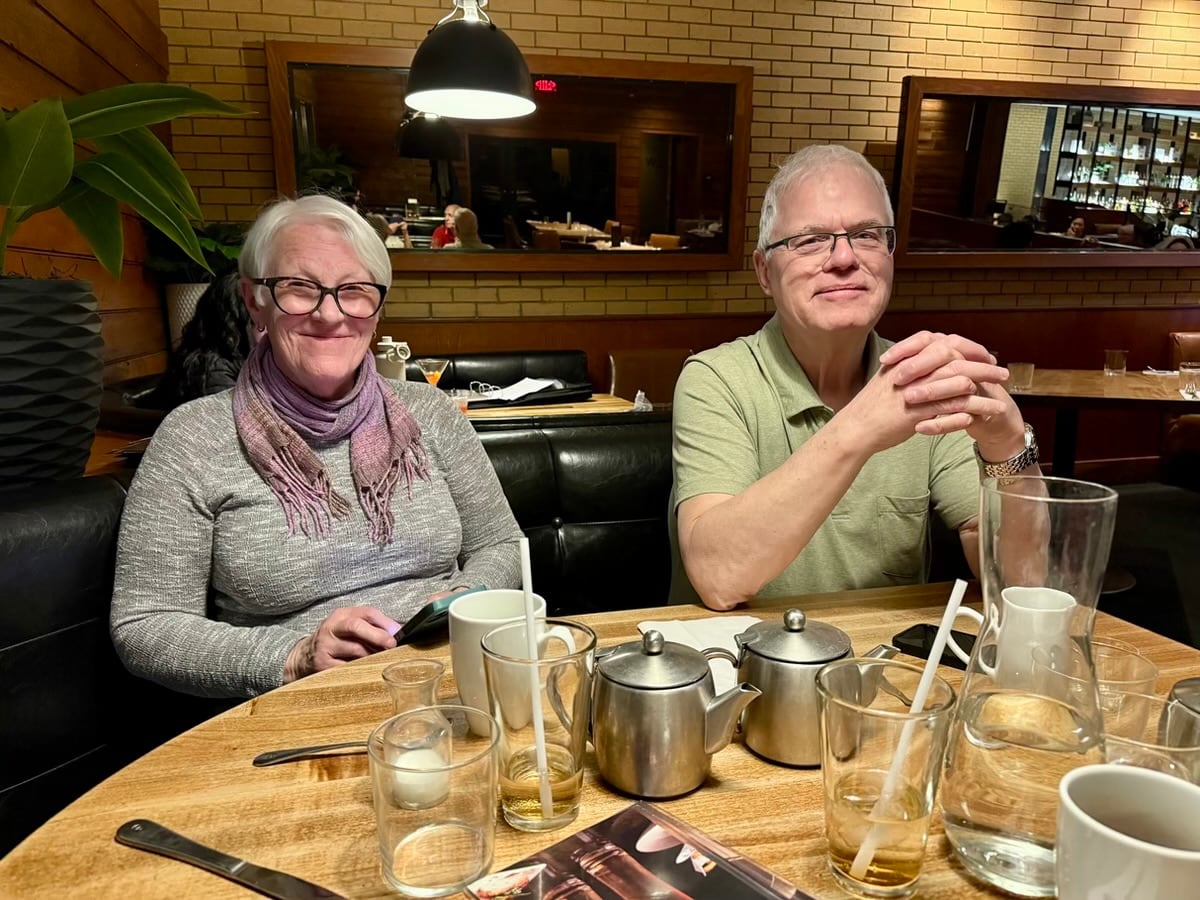 A man and a woman sitting in front of a wooden table with multiple glasses on it.