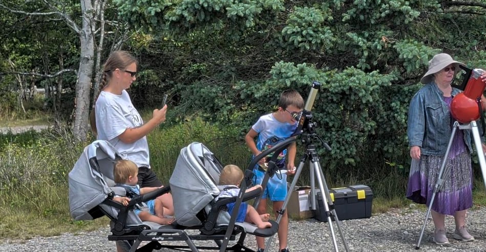 A young boy and elderly couple each standing at separate telescopes, outdoors on a sunny day.