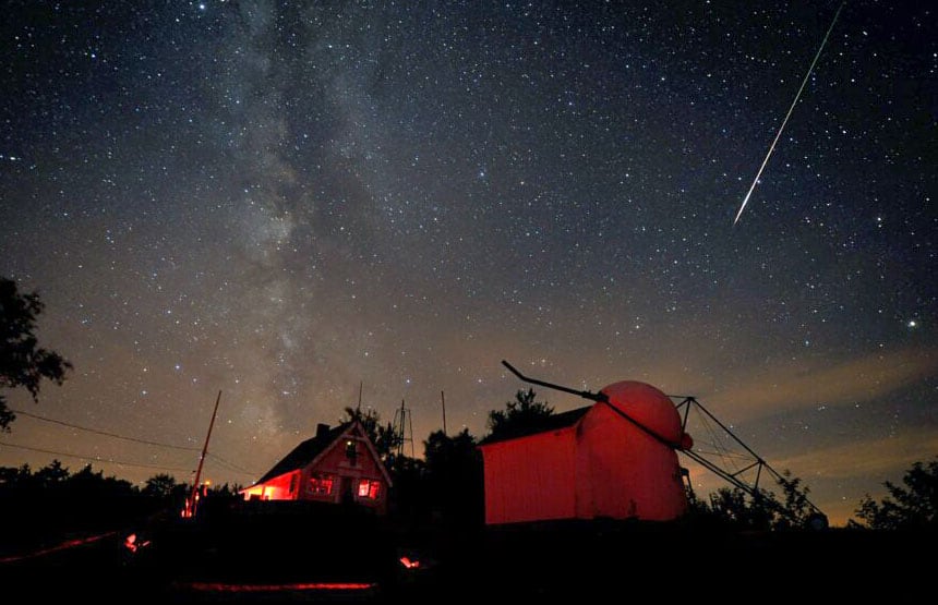 The Milky Way stretches over a home and a small observatory with a bright meteor streaking across the sky.
